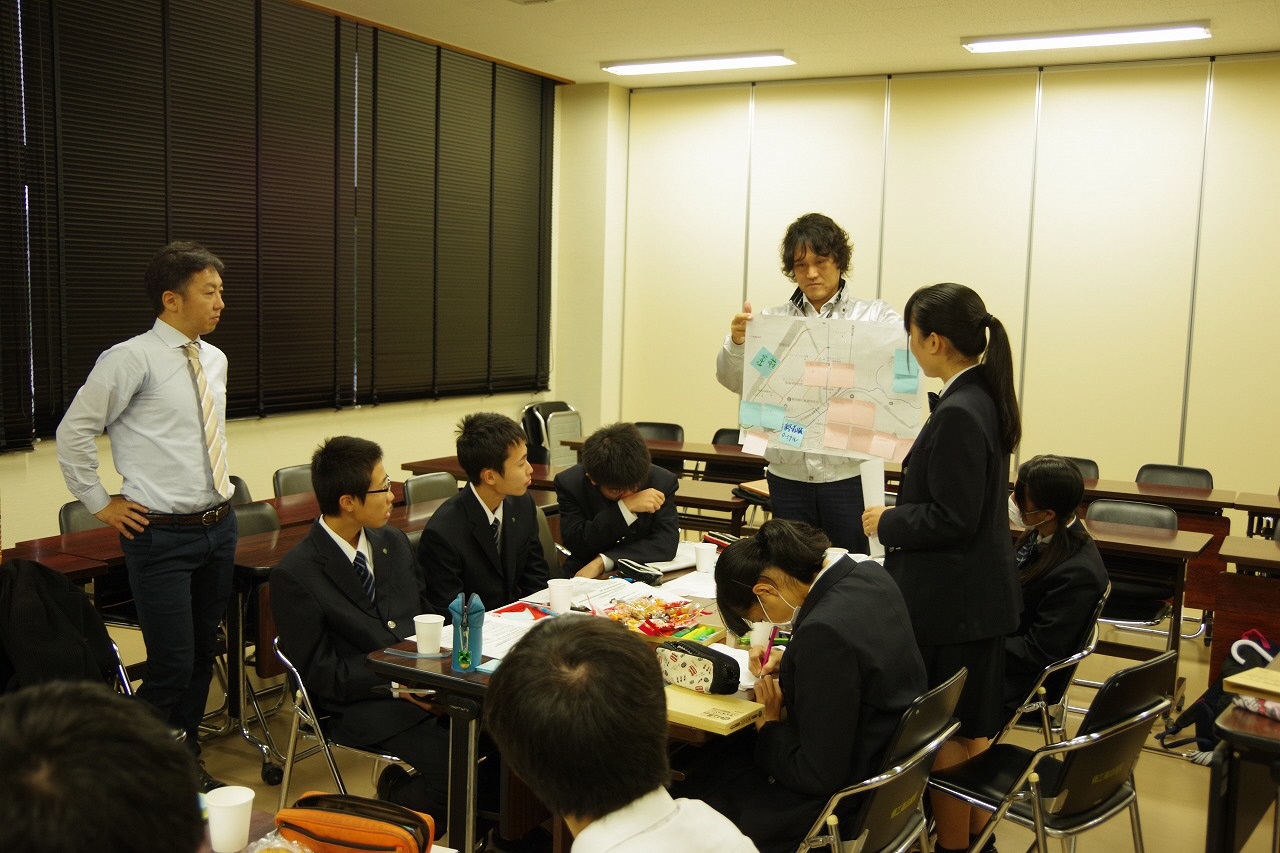 前方に立つ市職員が資料を持ち生徒の一人が立って発表し、それ以外の生徒は座って聞いている様子の写真