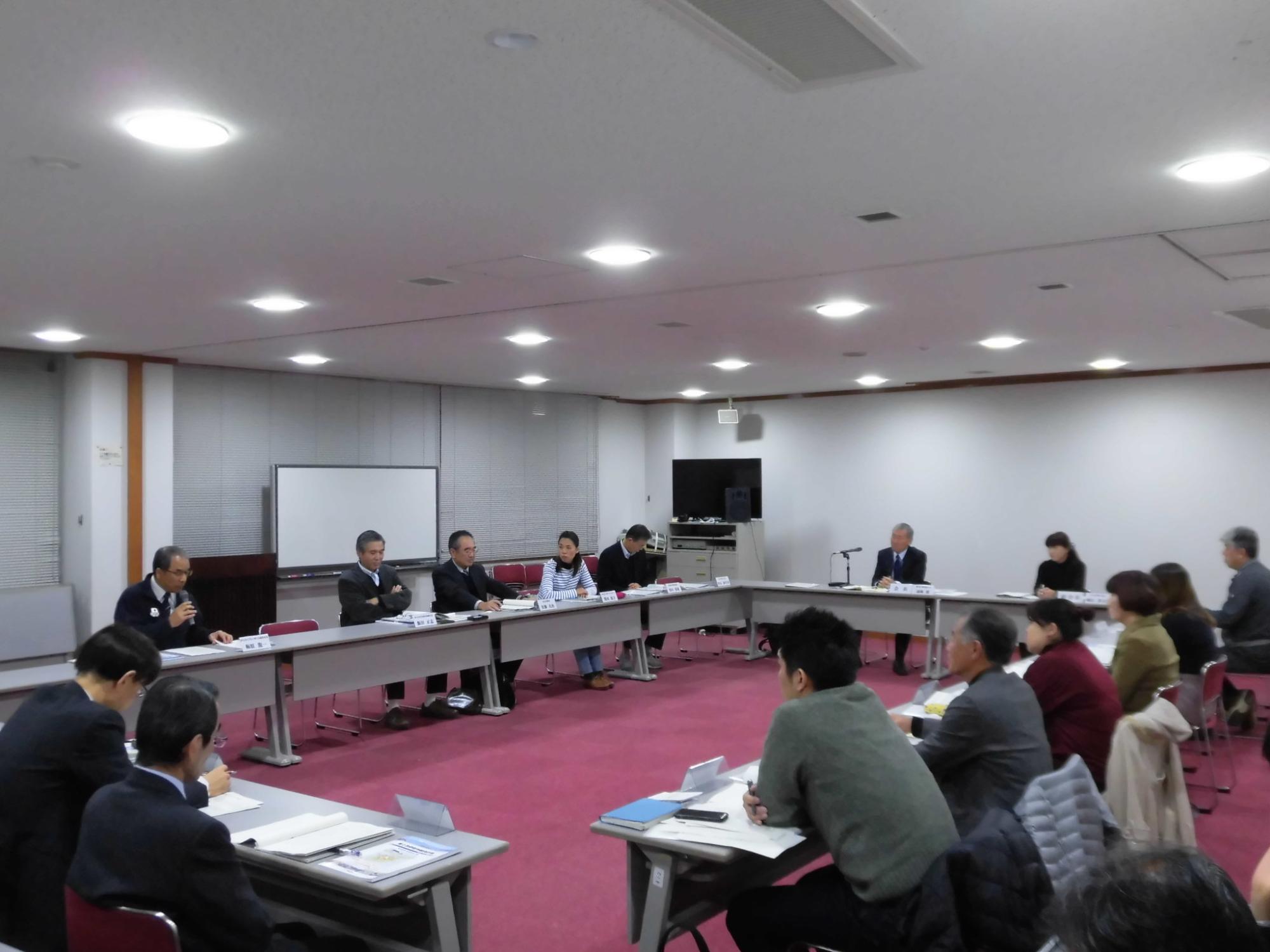 内側に向かい合うように長方形に並べられた席に座り、マイクをもって発言する男性の話を聞いている様子の写真