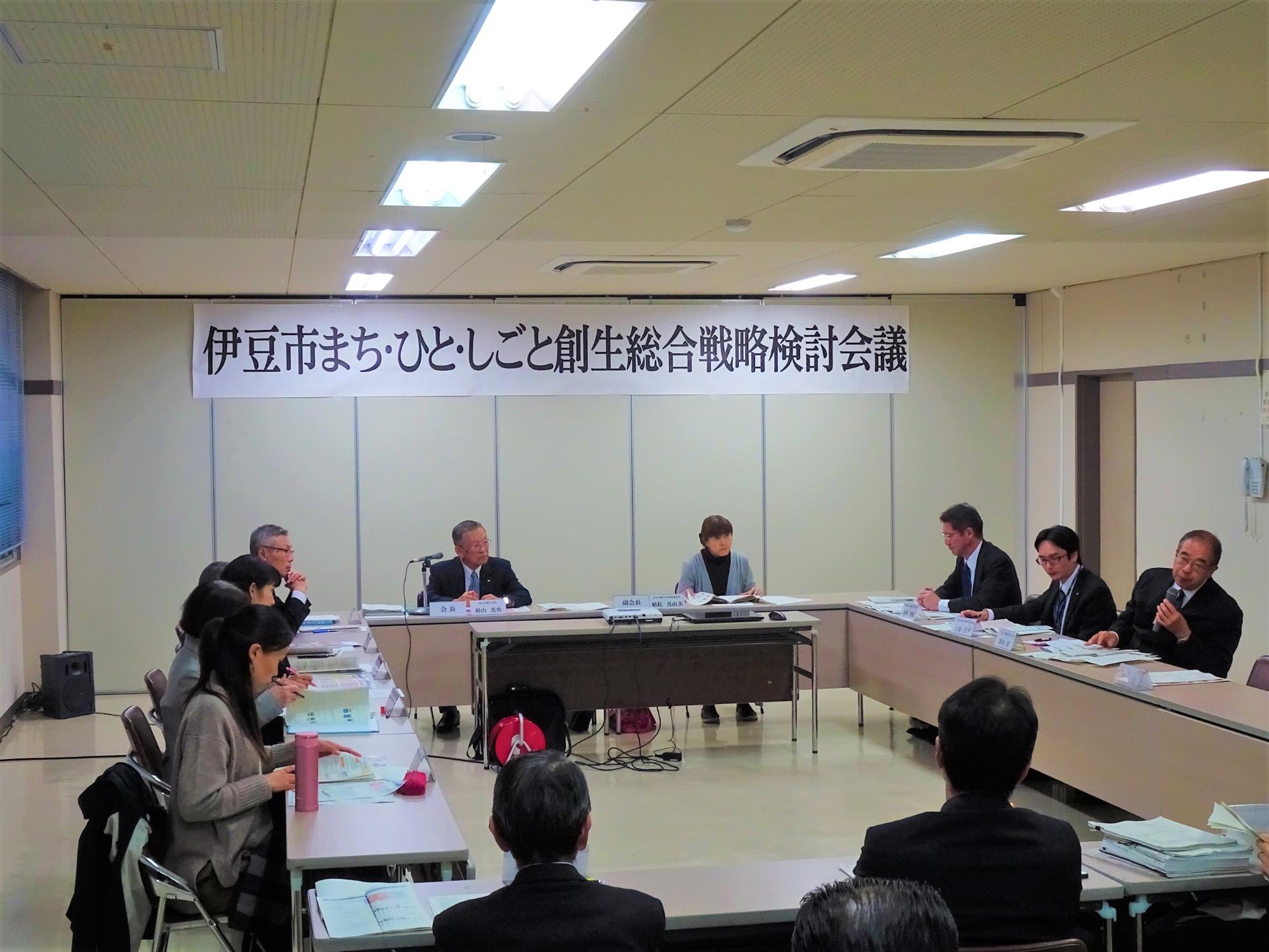 長方形に並べられた席に座った参加者が、右側の席に座りマイクを持って話をしている男性の話を聞いている様子の写真