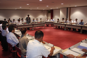 広い会議室で全員が内側を向く形で四角に机が置かれ、四角の中に置かれた長机の前に立つ男性がマイクをもって発言する様子の写真