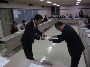 内側に向かい合うように長方形に机が並べられた会議室内で、市長が杉山会長に諮問書を手渡している様子の写真