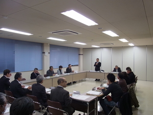内側に向かい合うように長方形に並べられた席に座り、1人の男性が立ってマイクを持ち発言をしている様子の写真