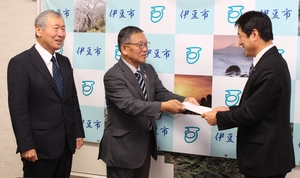 伊豆市の会見パネルの前で遠藤副会長の隣に立つ杉山会長が市長に答申書を手渡している様子の写真
