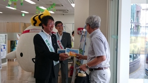 タスキを付けた市長と野村常務が男性にパンフレットを渡している様子の写真