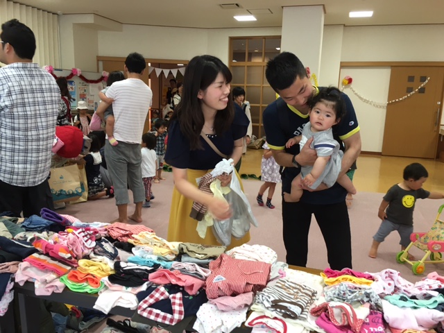 テーブルの上にたくさんの子供服が並べられている子供服お下がり会のブースにいる子供服を持った女性と赤ちゃんを抱っこした男性の写真