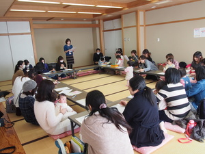 内側に向かい合うように長方形に並べられた席に座り、1人の女性が立って資料を見ながら発言をしている様子の写真
