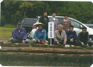 花の苗が植えられた木枠の花壇の後ろに、男女が2列に並んでいる記念写真