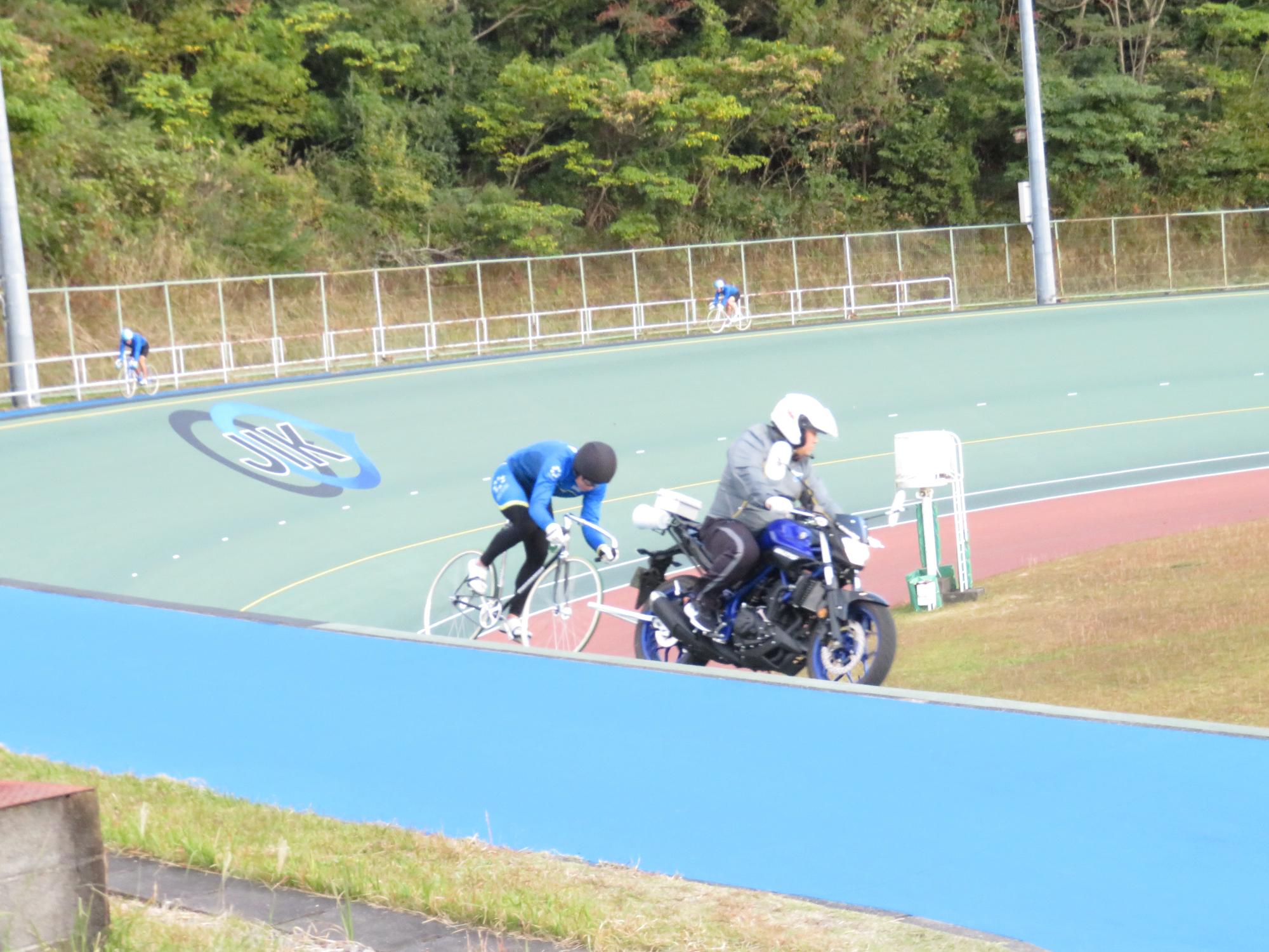 屋外のトラックを一台のバイクと競輪用の自転車が走っている写真