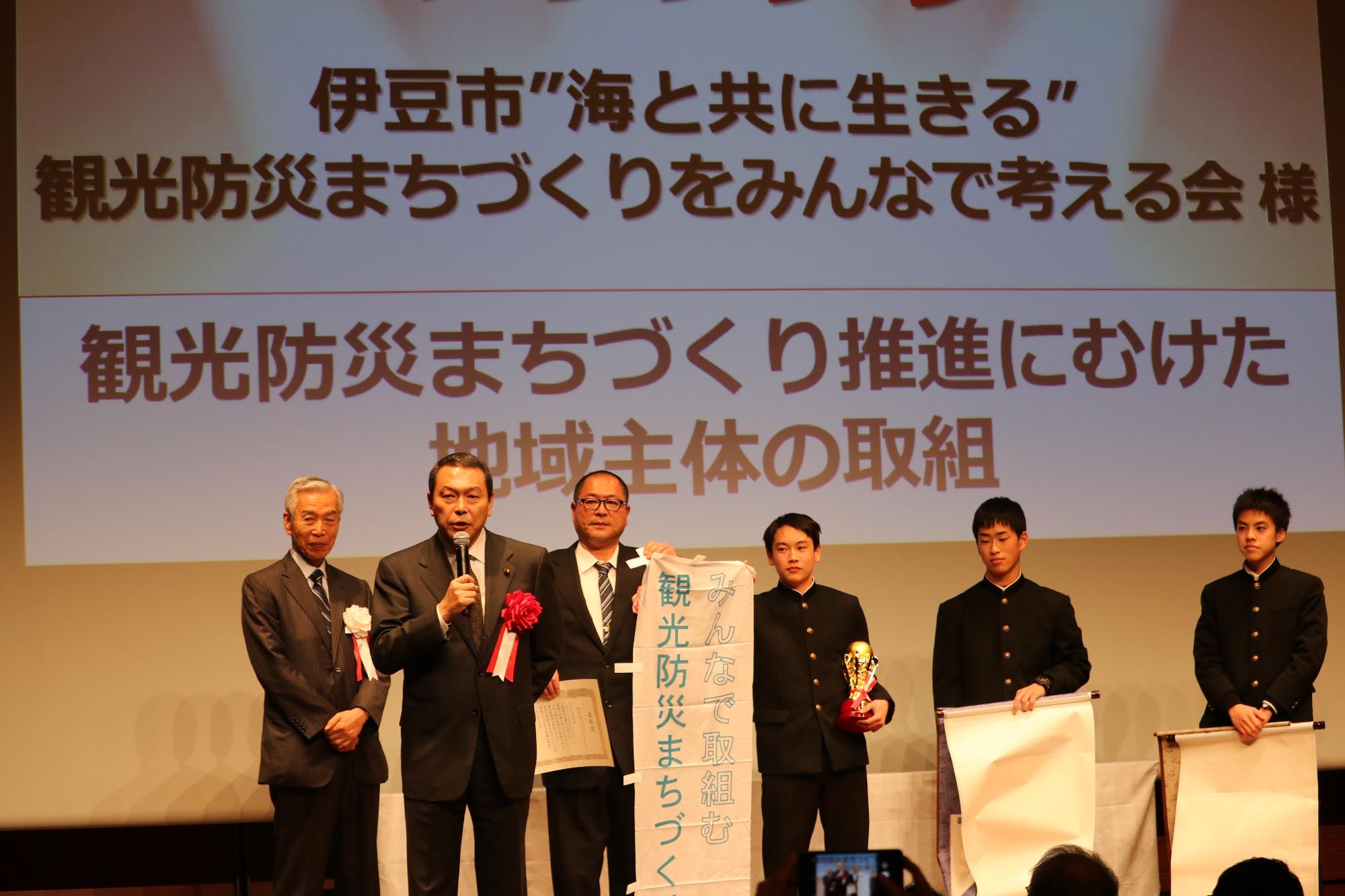 受賞された方々の前で小此木国土強靭化担当大臣がマイクを持ち話をしている表彰式の様子の写真