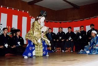 仮面をつけ金色の着物を着た男性が扇子を持ちながら踊っていて、周りを紺色の着物を着た祭囃子の男性達が囲んでいる写真