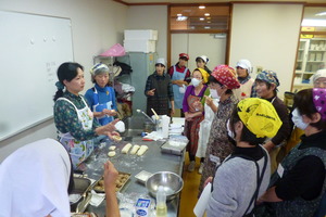 肉まんの包み方を実演しながら説明する女性と、周りに集まり説明を聞いている参加者の方々の写真