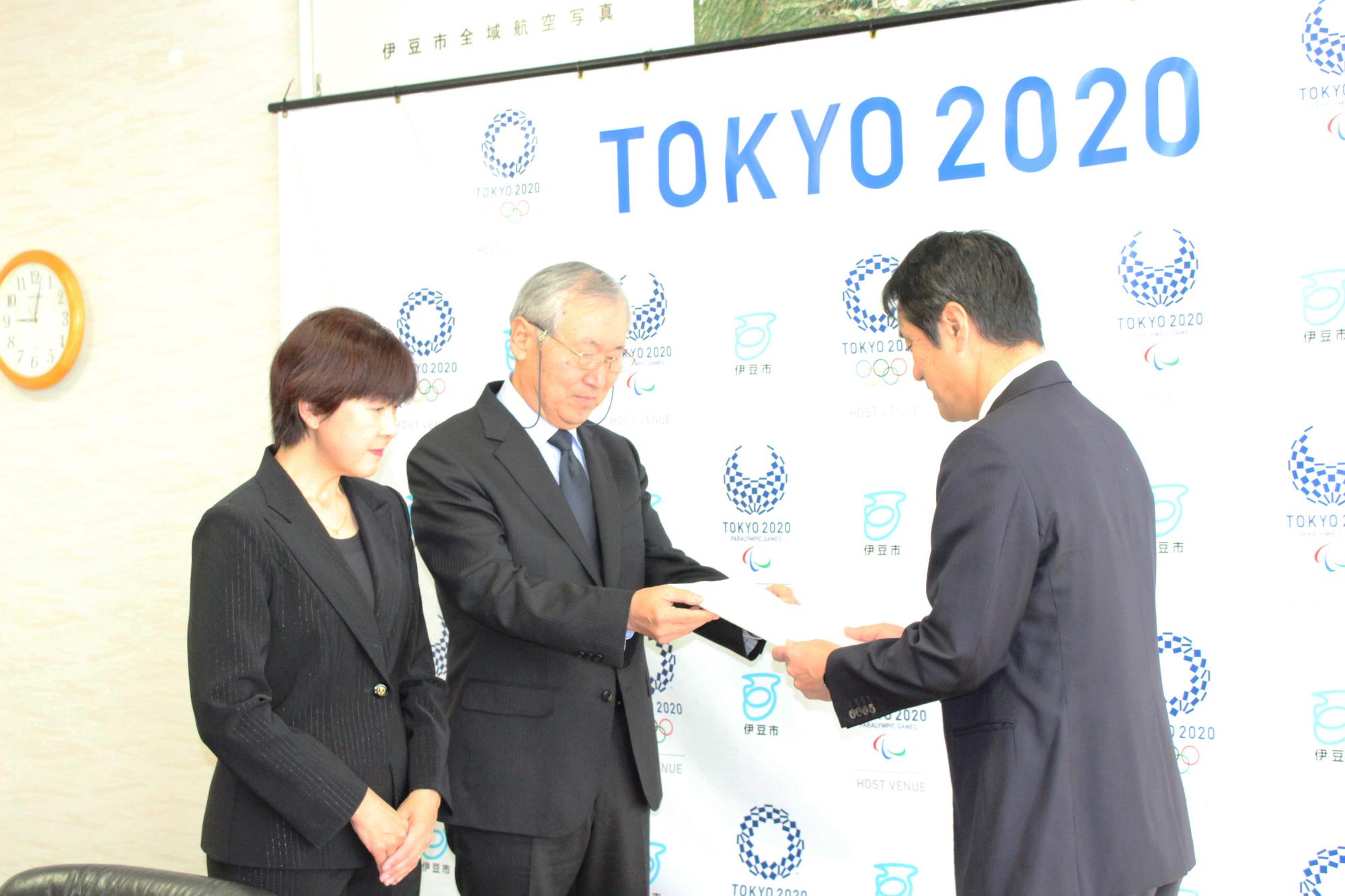 TOKYO2020の会見パネルの前で遠藤会長と植松副会長が並んで立ち、遠藤会長が市長に答申書を渡している様子の写真