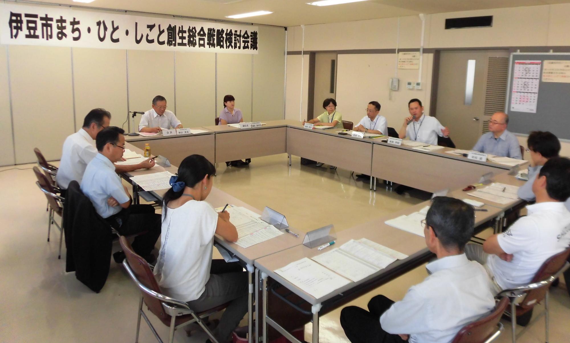 長方形に並べられた席に座った参加者が、右側の席に座りマイクを持って話をしている男性の話を聞いている様子の写真