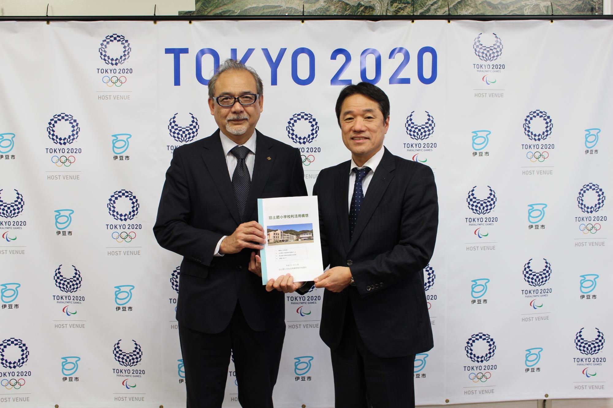TOKYO2020の会見パネルの前で活用構想をまとめた資料を持つ作山会長と菊池市長の写真