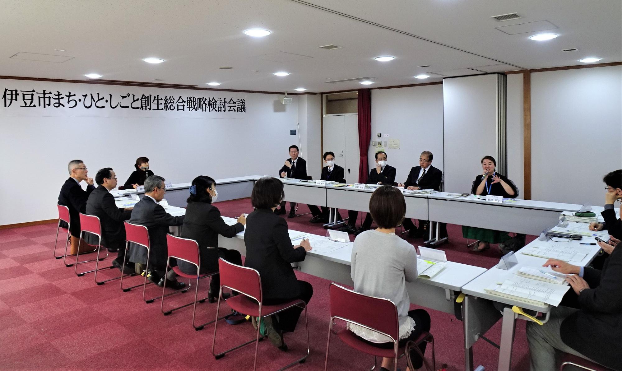 長方形に並べられた席に参加者が座り、女性がマイクをもって話をしている様子の写真