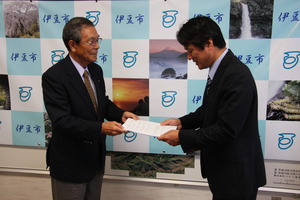 伊豆氏の会見パネルの前で遠藤会長が市長に答申書を手渡している様子の写真