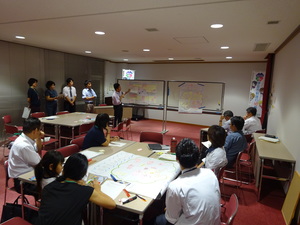 会議室前方で2つのホワイトボードを使って成果発表を行うグループとそれを聞いている参加者の方々の写真
