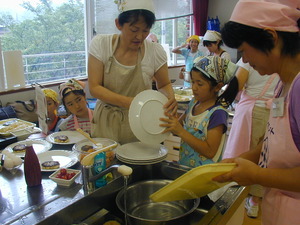 三角巾を頭に巻き、エプロンをつけた親子達がお皿を洗ったりなどの作業をしている写真