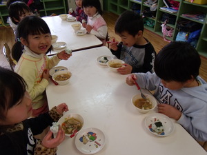 子ども達がテーブルに座って豚汁を食べていて、一人の女の子がカメラに向かってほほ笑んでいる写真