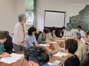 長机に参加者達が資料を見ながら座っていて、手前の白い上着を着た女性が立って話をしている食育推進会議の写真