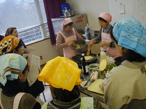 講師の女性2人を囲むように頭に三角巾を巻いてエプロンを着けた参加者の女性たちが立っている写真