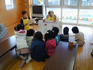女性が大きな紙芝居を読んでいて、紙芝居を囲むように子供たちが集まっている写真