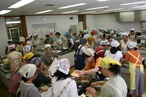 調理台の周りを囲むように三角巾を頭に巻いた参加者達が集まり作業をしている写真