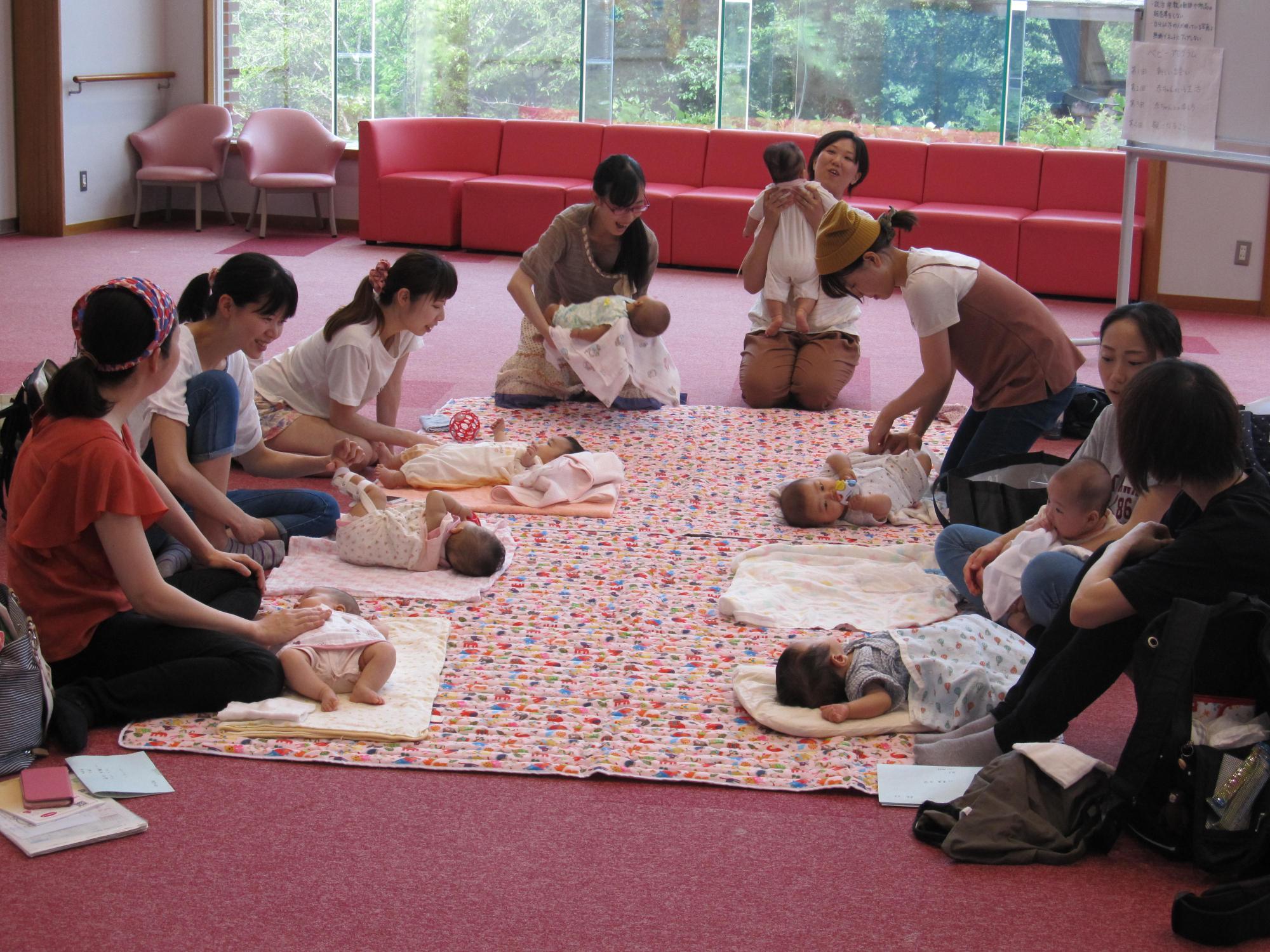 8組の母子が床に敷かれた柔らかい布のマットの上に赤ちゃんを寝かせ、赤ちゃんの様子を見たり母親同士で話し合ったりしている様子の写真