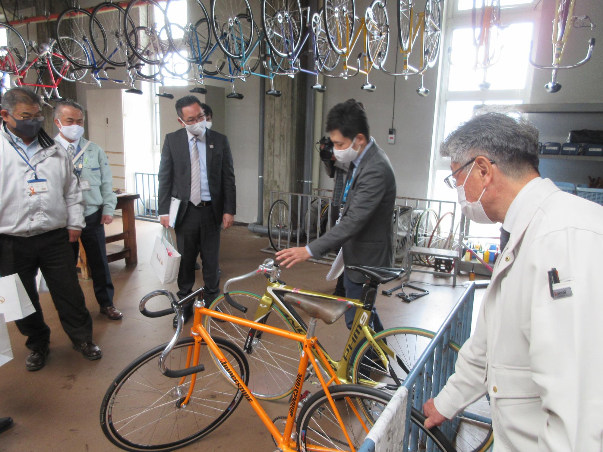 天井に自転車が並べて吊り下げられている実習場で職員達が2台の競輪用自転車を見学している写真