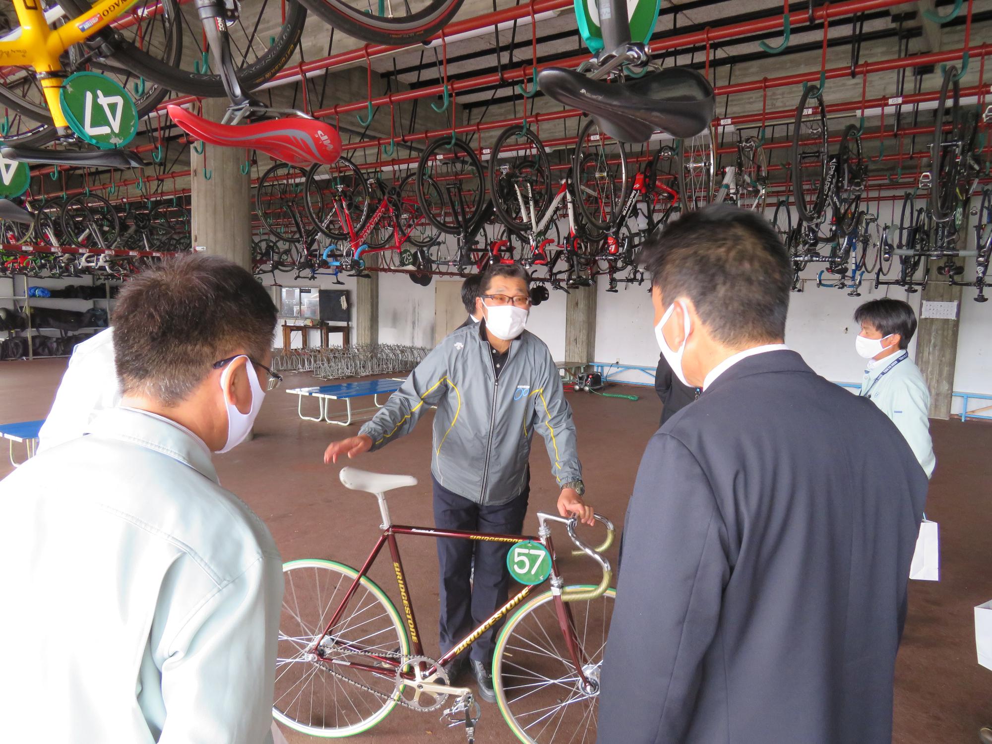 格納庫でメガネをかけた男性が57番と番号のついた自転車を触りながら職員達に説明をしている写真