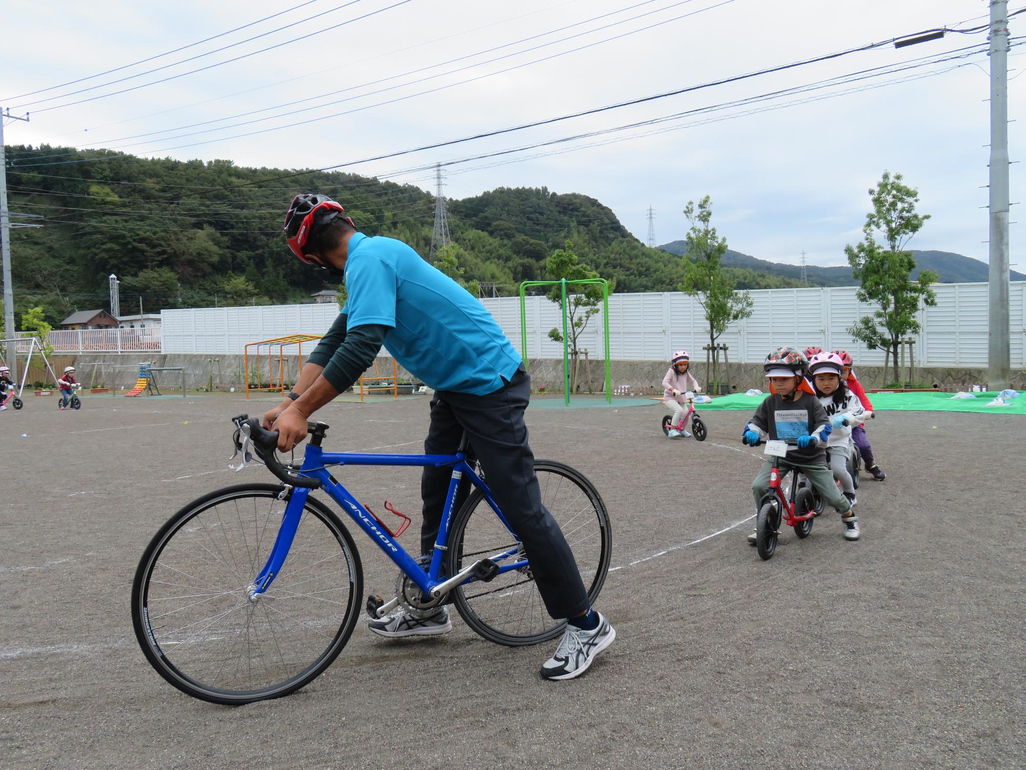ヘルメットを被って自転車にのった子供たちが縦に並びながら、先頭にいる自転車に乗った男性の後をついて走っている写真