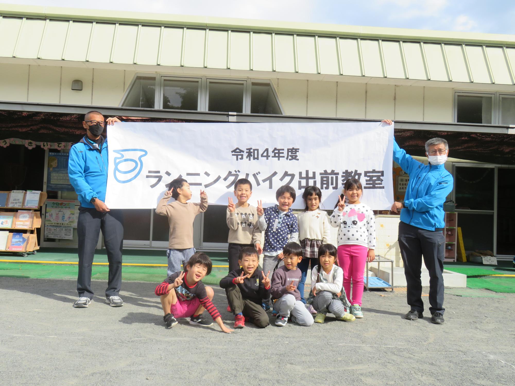 令和4年度ランニングバイク教室と書かれた幕の前で熊坂こども園の9名の子供たちが並んでいる集合写真