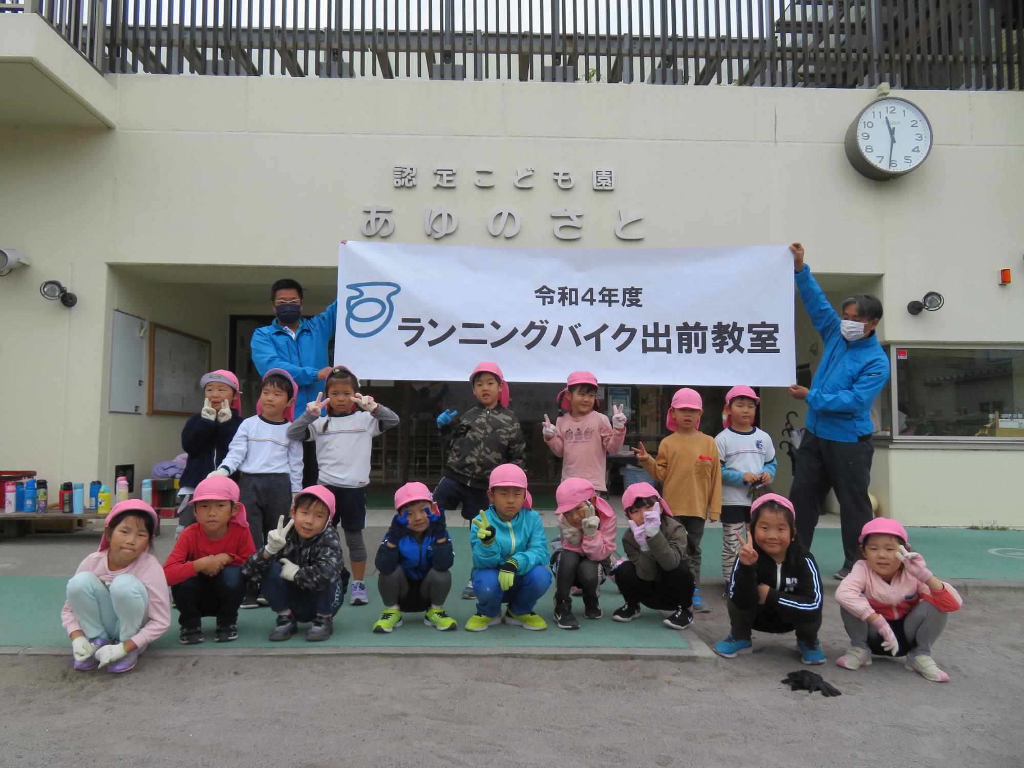 令和4年度ランニングバイク教室と書かれた幕の前でピンクの帽子を被った認定こども園あゆのさとの子供たちが並んでいる集合写真