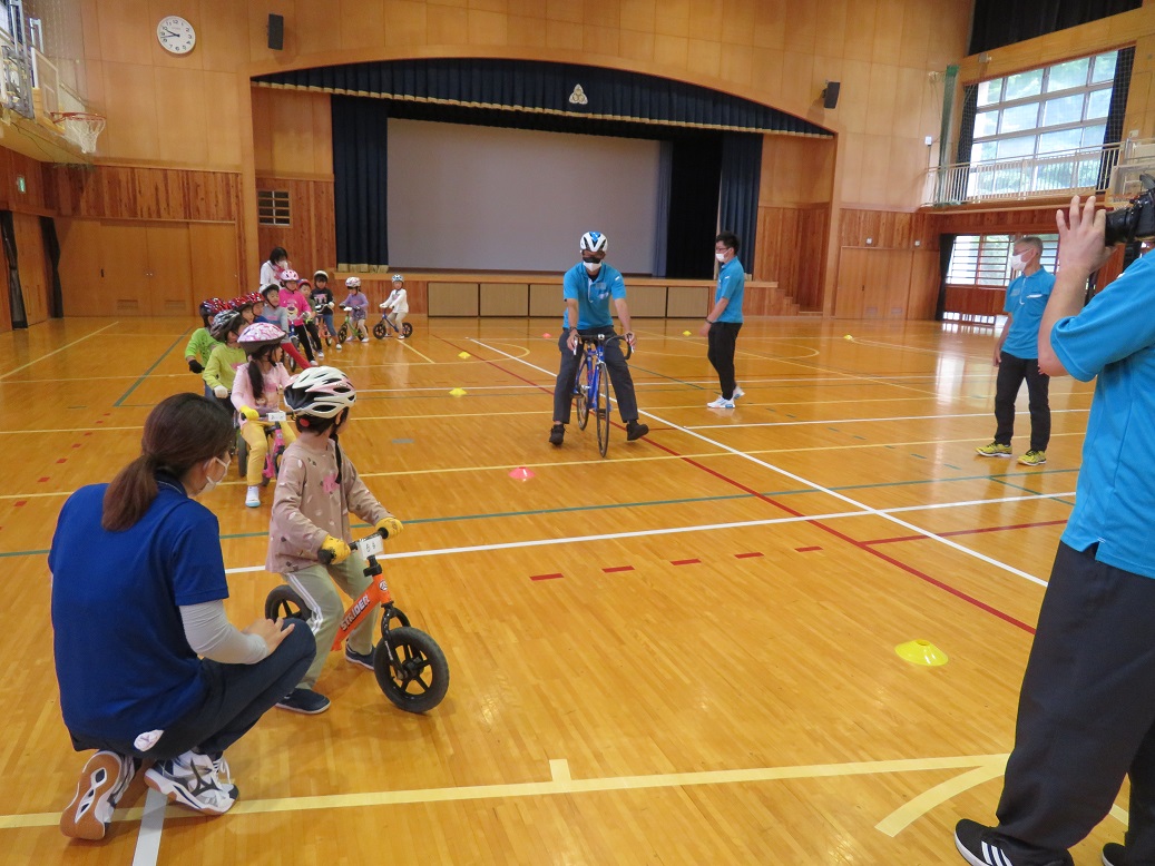 ヘルメットを被った子供たちが自転車にまたがり、中央にいる自転車に乗った男性の話を聞いている写真