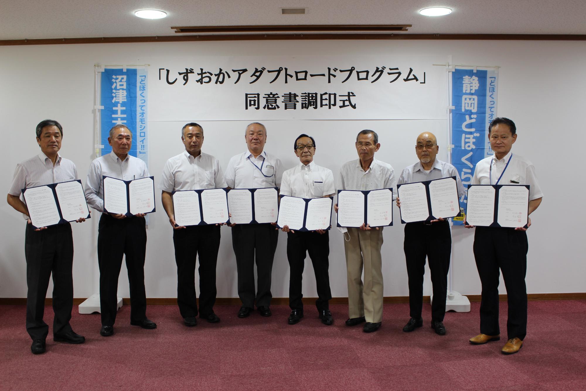 男性8名が横一列に並び、6団体が同意書を持って記念撮影をしている写真