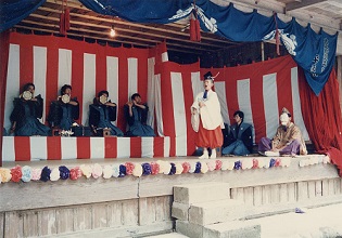 紅白の幕がかかった舞台上で着物を着た男性達が並んで堤太鼓を叩いていて、中央に白い衣装の男性が立っている写真