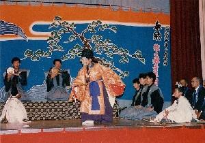 松の木などが描かれた背景幕の前で裃を着た男性達が正座で小鼓を叩きオレンジ色の束帯を着た男性が舞を披露している写真