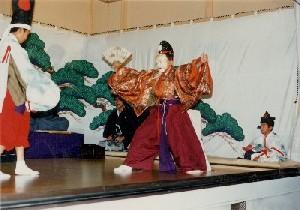仮面をつけ着物を着た男性が舞台上で扇子を持ちながら両足を広げ立っている写真