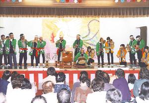 紅白の幕がかかった舞台上で緑の法被を着た男性達が和太鼓や笛などを演奏している写真