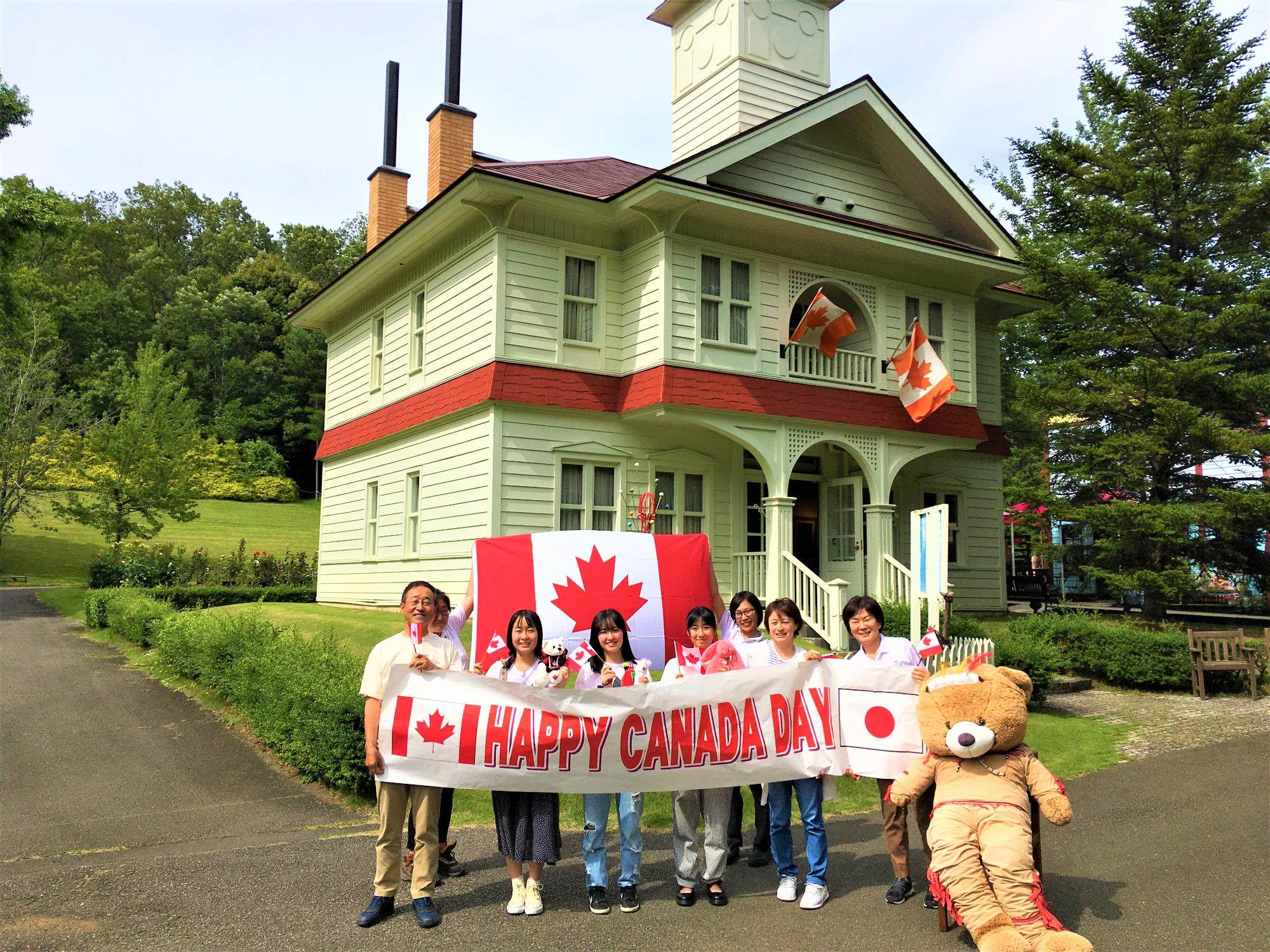 カナダと日本の国旗の間に「HAPPY CANADA DAY」と書かれた横断幕を持った8人の方の集合写真