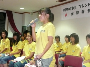 お揃いの黄色いTシャツを着た生徒たちが並んで座り、1人の女子生徒が立ってマイクを持ち話しをしている様子の写真