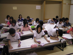 体操服やジャージを着た子供たちが、白い大きな紙に油性ペンで英語で何かを書いている様子の写真
