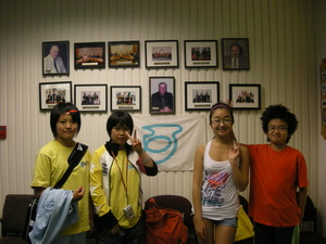 額に入れられた11枚の写真と伊豆市の市旗が飾られている前に立つ4人の子供たちの写真