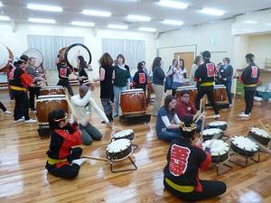 法被を着た伊豆総合高校郷土芸能部の部員が、外国人男女に太鼓を教えている写真