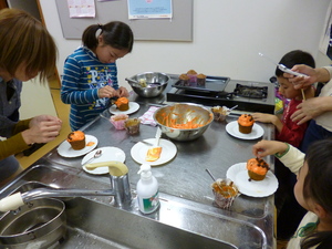 女性や、子供たちがカップケーキにオレンジ色のアイシング等ジャックランタン風の飾りつけをしている写真