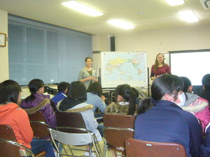 世界地図の横に立ち講義をするkimさんとご友人と、椅子に座って話を聞いている生徒たちの写真