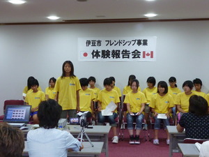 お揃いの黄色いTシャツを着た生徒たちが2列に並んで座り前列の女の子が立って発表をしている様子の写真