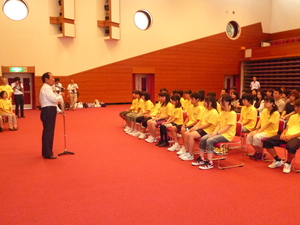お揃いの黄色いTシャツを着た生徒たちが並んで座り、マイクの前立つ石渡会長の話を聞いている様子の写真