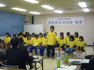 お揃いの黄色いTシャツを着た生徒たちが2列に並んで座り前列の男の子が立って発表をしている様子の写真