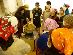 大きな鍋でカレーを煮込んでいるインド人の女性と、周りに集まりそれを見ている参加者の方々の写真